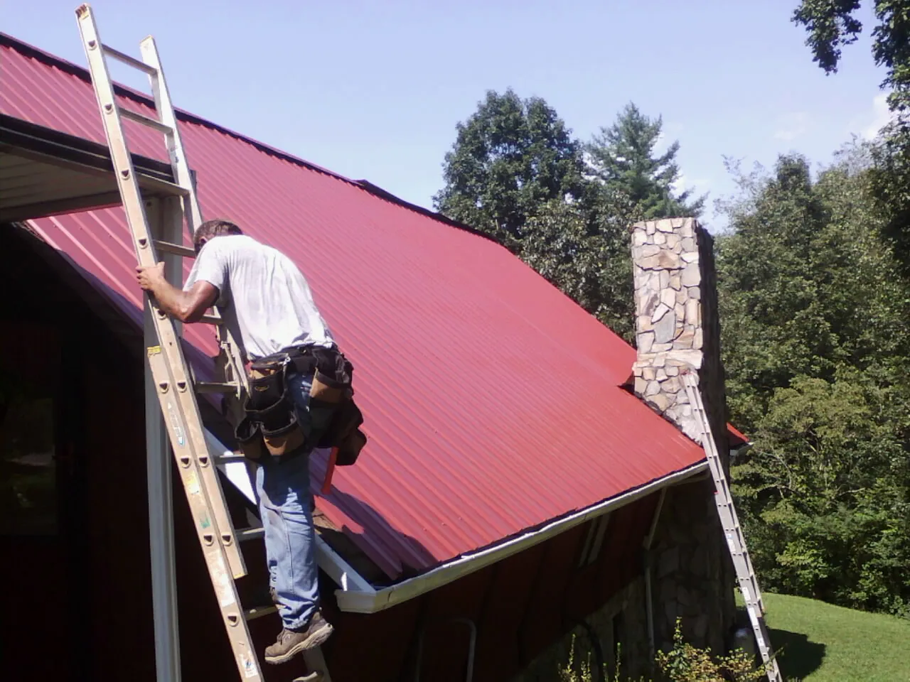 Roofer on a ladder beside a red metal roof and stone chimney — Soffit & Fascia Repair by Roofing Brothers in Du Pont